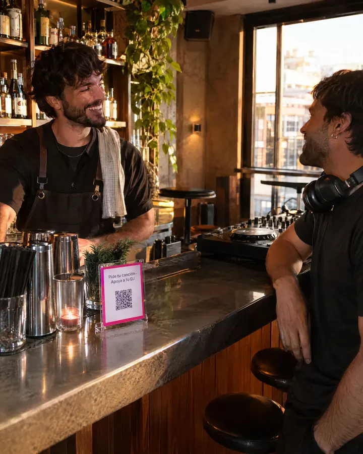 Antes de abrir, un camarero y un DJ charlan en la barra junto a una tarjeta con el QR «Pide tu canción · Apoya a tu DJ».