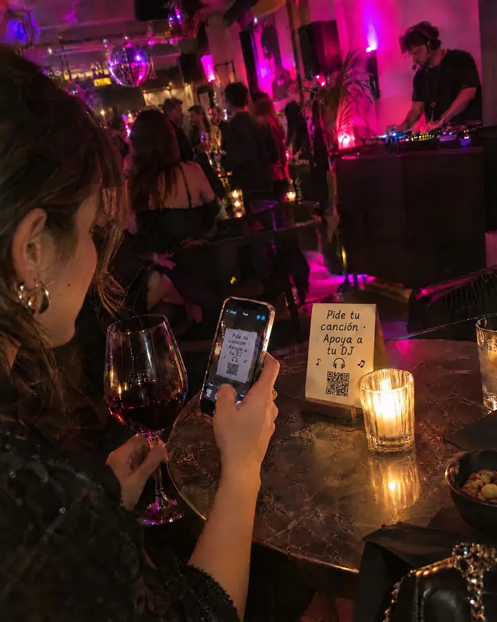 Una coctelería con un DJ en la esquina y una clienta escaneando el QR con el móvil.
