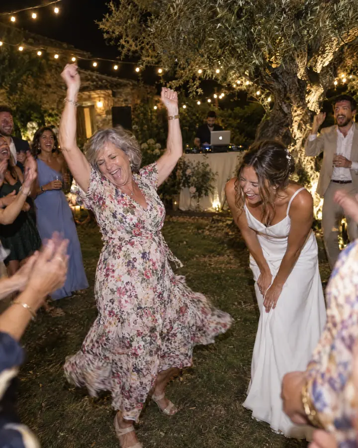 At a farmhouse garden wedding, a grandmother dances with her arms up while the bride doubles over laughing beside her.