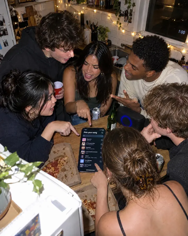 At a house party, six friends crowd around a phone in the kitchen showing the "Pick your song" screen.