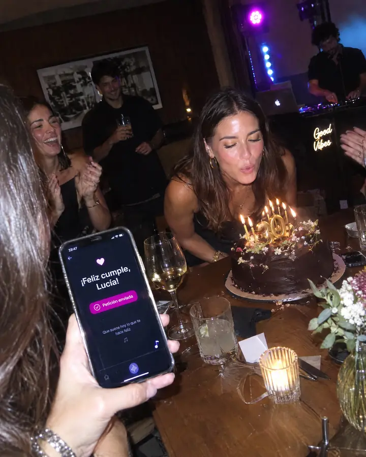 En un cumpleaños, una amiga enseña en el móvil la dedicatoria «¡Feliz cumple, Lucía!» mientras ella sopla las velas.