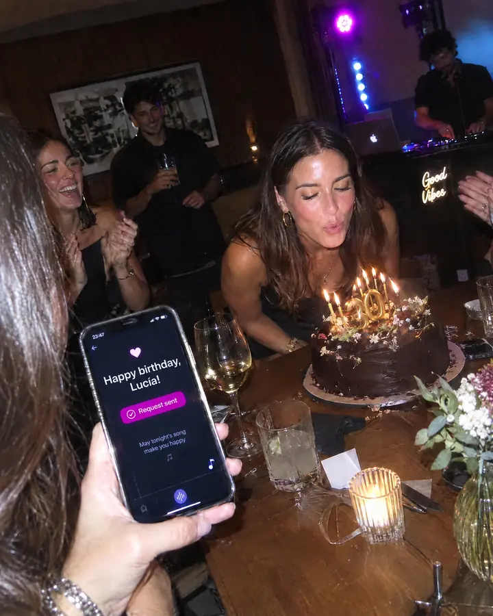 At a birthday, a friend shows the dedication "Happy birthday, Lucía!" on her phone while Lucía blows out the candles.