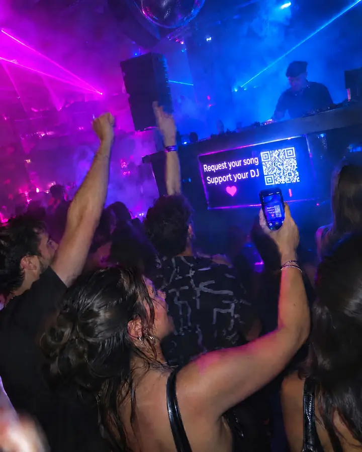 A crowded club dance floor with the DJ booth behind and phones in the air.