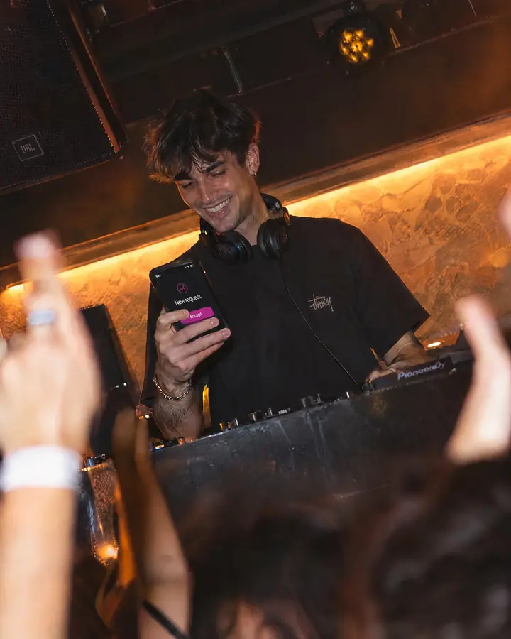 A DJ in the booth, one hand on the mixer, checks a WeSound song request on a phone.