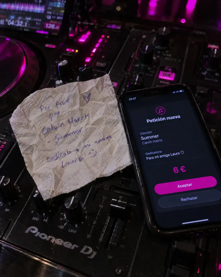 Sobre la mesa de mezclas, una servilleta arrugada con una petición escrita a boli junto a un móvil con la petición de WeSound ordenada: canción, dedicatoria y 6 €.