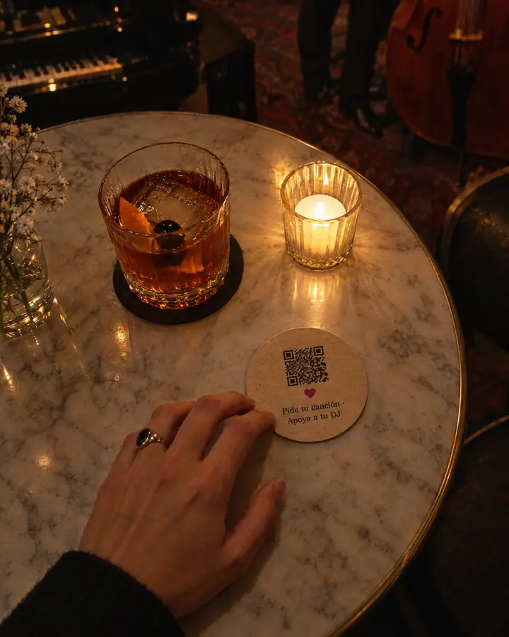 Vista desde arriba de una mesa de un bar de jazz: un cóctel, una vela y un posavasos con el QR y «Pide tu canción · Apoya a tu DJ».
