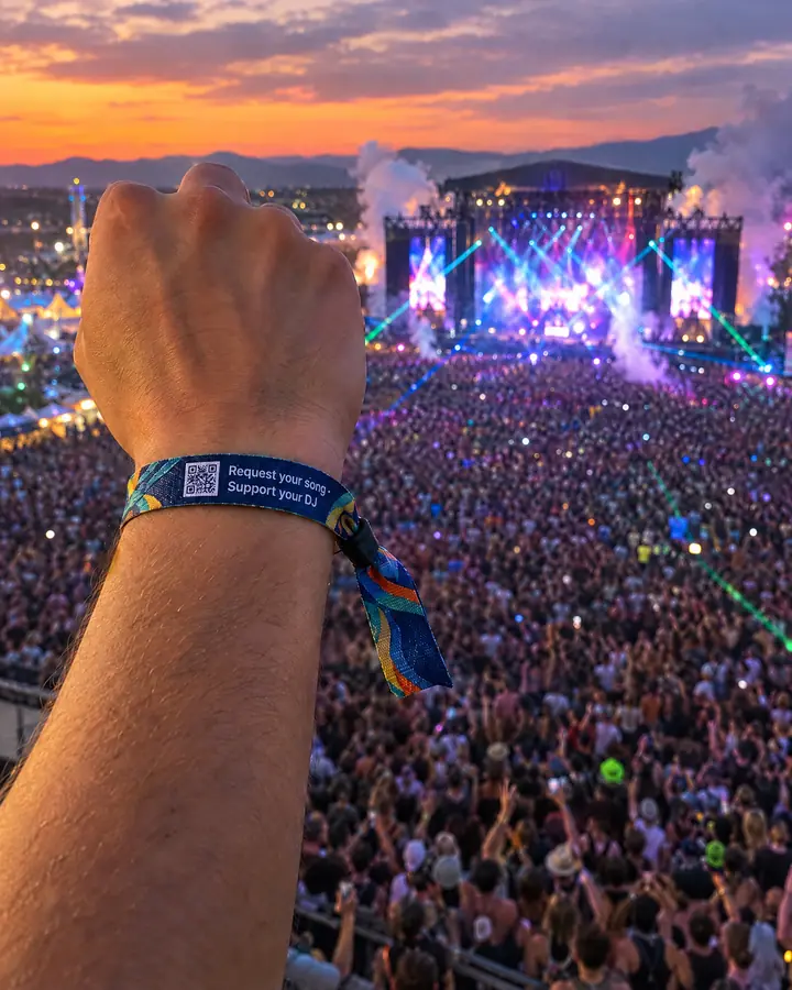At a big festival at sunset, a raised arm wearing a fabric wristband with the QR code and "Request your song · Support your DJ".