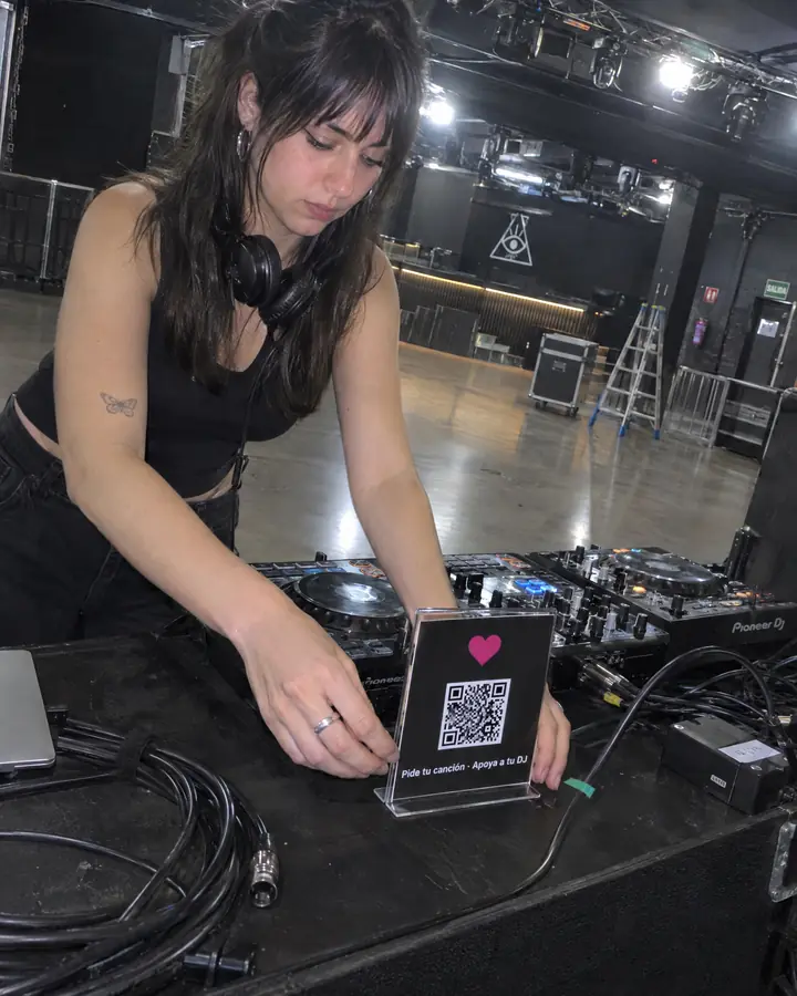 Una DJ coloca en la cabina, antes de abrir, un soporte con su QR y el mensaje «Pide tu canción · Apoya a tu DJ».