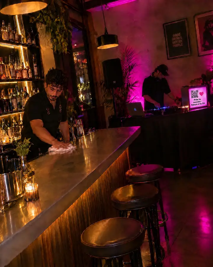 Un bar de copas con luz baja y el DJ en la cabina; en una pantalla junto a la cabina se ve el código QR del DJ.