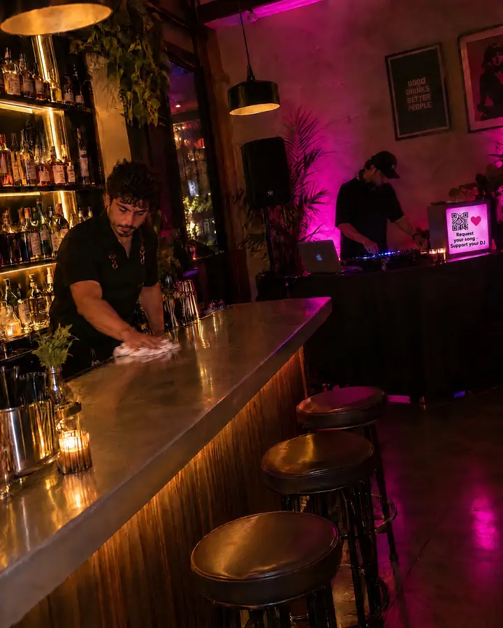 A dimly lit cocktail bar with the DJ in the booth; a screen next to the booth shows the DJ's QR code.